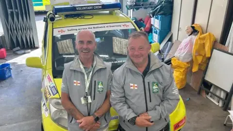 Two men stand in front of a bright yellow small car, with sirens fitted and sponsorship stickers. They are standing in a warehouse, with outboard motor, and a crash test dummy. They are wearing pale grey uniforms of Guernsey's St John volunteers, and smiling. 