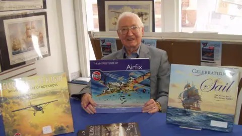 Anthony Cross A grey haired man, wearing steel-rimmed glasses and a grey jacket, at a table. He is hold a blue book called "THE VINTAGE YEARS OF Airfix". To the man's right is a yellow book entitled "CELEBRATION OF LIGHT - THE AVIATION ART OF ROY CROSS. To his left is a book called "CELEBRATION OF Sail - The Marine Art of Roy Cross RSMA" 