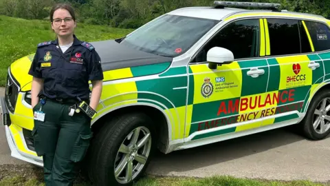 Hannah Whitelegg, standing in front of an ambulance emergency response vehicle, that has yellow and green livery. Hannah is wearing dark green trousers, and a blue top, an responder uniform, with her hands in her pocket.