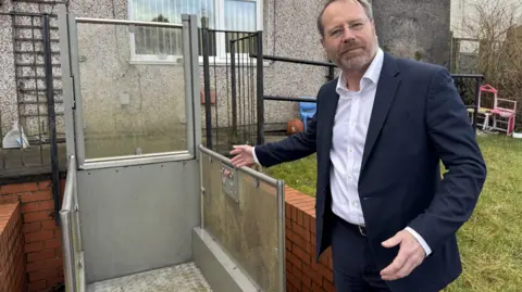 An image of the Rossendale and Darwen MP standing next to the lift in Millie Mulcahy's garden. It is a metal lift suitable for a wheelchair and is surrounded by a red brick wall. Andy McNae MP is wearing a navy suit with a white open necked shirt, he is pointing at the lift. There is a lawn behind him with children's toys in the background.