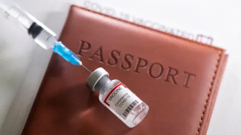 A syringe inserted into a bottled labelled Covid-10 vaccine. They are lying on a passport in a brown leather case.