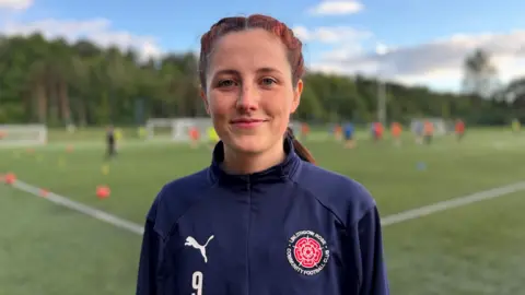 A woman with dark hair, tinted red, wearing a tracksuit and standing by the side of a football pitch 