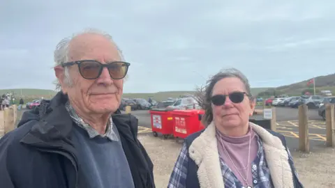 George Carden/BBC A man and a woman are standing near the car park wearing winter coats and sunglasses. 
