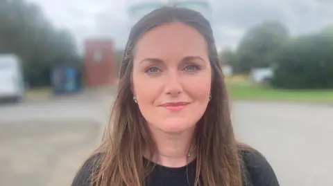A woman with long brown hair smiles at the camera. She is wearing a black top and a necklace. Behind her are a number of trees.