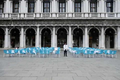 Reuters A waiter stands by empty tables outside a restaurant at St Mark's Square after Italy's government adopted a decree with emergency new measures to contain the coronavirus, in Venice. March 5, 2020.