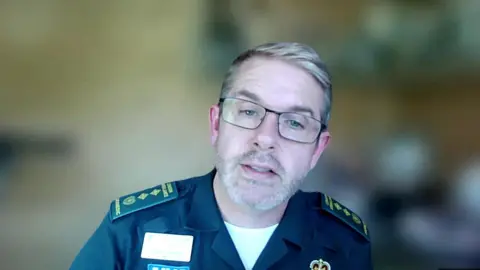 A middle aged man with glasses and a greying beard speaks on a Zoom call. He is wearing a London Ambulance Service uniform. 