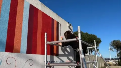 A wall with blue, dark red and orange stripes on it. There is a person on scaffolding adding another stipe at the end. In the background is a blue sky and trees. 