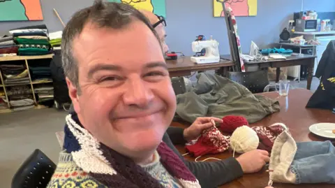 Mousumi Bakshi/BBC Adam Cleevely is on the left of the image and is smiling. He has short brown hair and is wearing a patterned jumper. Behind him is a table with a white sewing machine on it and fabrics. 