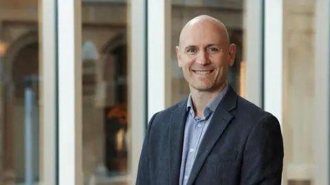 Tourism NI George Diamondis, a man with dark jacket and light blue shirt, who is smiling. He is standing in front of a series of windows.