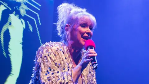 Cheryl Baker in a white shirt with silver circles on it. She is singing into a microphone with a red foam top.