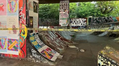 M32 Skatepark in Eastville, Bristol. Graffiti and posters can be seen all over the pillars and walls. Yellow police crime scene tape can be seen across the photo. 