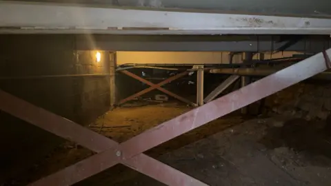 A view of the theatre basement with steel girders, bare brick walls and what appears to be an old seawall to the right. The space is dimly lit by a yellow light.