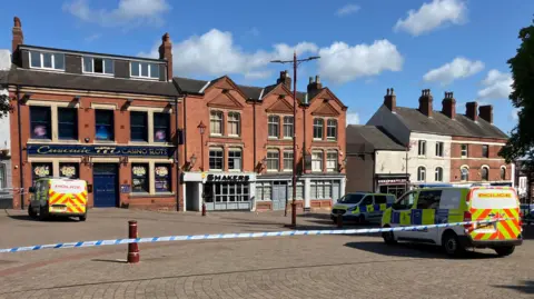Police tape across a large area of an open pedestrianised square, with police vehicles parked up inside near to a row of pubs