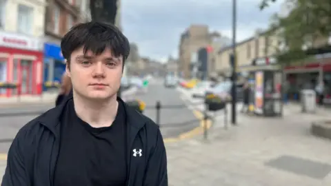A photo of Alex standing on a high street. He is wearing a black t-shirt and a black tracksuit. He had black hair and a eyebrow stud