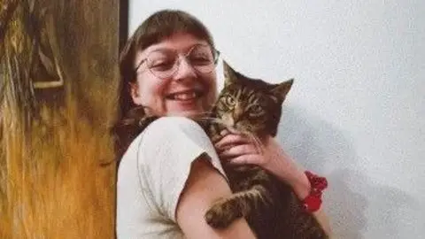 Paul Burke Picture of Becky Burke cradling a tortoiseshell tabby cat in her arms. She grins at the camera and wears thin round glasses, with brown short hair and a full fringe. She wears cream jeans with a wide brown belt and a cream t-shirt, a tattoo can be seen on her bicep of three line-drawn tulips. 