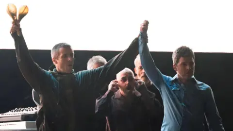 Liam and Noel hold their hands up in the air on stage 