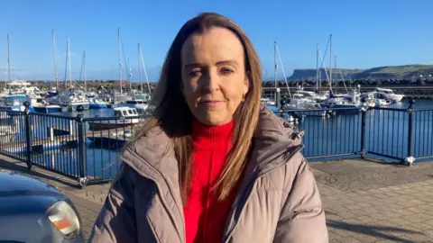 Councillor Cara McShane is wearing a bright red top and light purple coat. She is looking right at the camera and has long brown hair. She is standing in front of the marina and boats are docked. 