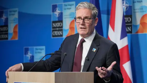 Keir Starmer gestures with his hand while speaking at a Nato summit on Wednesday in the Hague, he's wearing a suit and tie and glasses and stands at a podium with the UK flag in the background against a blue wall with the Nato logo on.