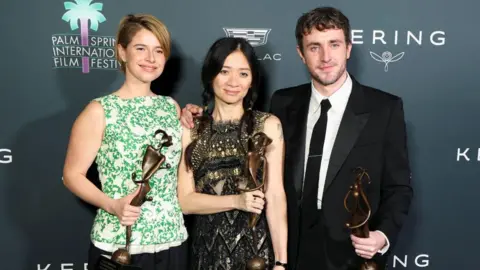 Reuters Jessie Buckley, Chloe Zhao and Paul Mescal stand with their arms around each other against a black backdrop for the Palms Springs International Film Festival. Buckley is wearing a white and green patterned top and is holding a bronze award. Zhao is wearing a black lace dress and also holding a bronze statue. Mescal on the right is wearing a black suit and tie with white shirt and is also holding a bronze statue award.