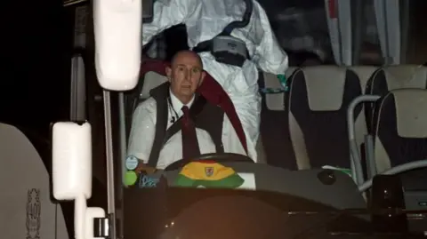 Andy Simonds sitting in the driving seat of the bus he drove, with a bucket Wales cap on the dashboard and people in PPE visible behind him