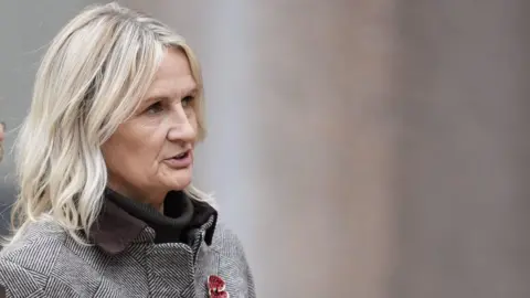 PA Media Lucy Ward is seen in conversation as she walks into court. She has short blonde hair and wears a grey coat with a red poppy badge.