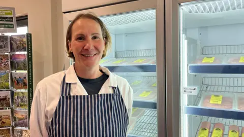 Ellen Knight/BBC Nicky Thomsett stands in front of two fridges in the farm shop, which have packets of sausages on their shelves. She is wearing a long white coat, with a dark blue top on underneath. Thomsett has a dark blue and white striped butcher's apron on over the top. She has brown, chin-length hair, and is smiling. 