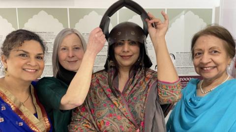 Sikh women in saris stand together, one is wearing a medieval helmet