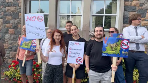 The image shows Emma Pang, on the left, standing amongst a group of protesters. She is a woman with long ginger hair, wearing a white t shirt and black-and-white baggy trousers. Next to her is a younger woman wearing black, and on the right is a man with a beard and moustache. All three are holding anti-GST placards. 