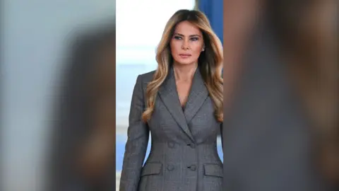 Melania Trump has long, styled, blonde hair. She is wearing a grey, double-breasted women's suit jacket.