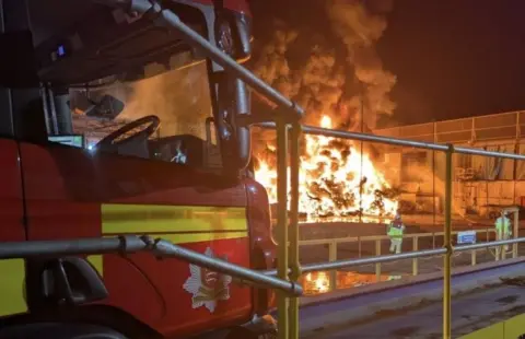 The fire burning at night with bright flames and a lot of smoke. A fire engine in the foreground and two fire fighters stand near the blaze.