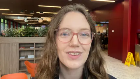 Jess Hindley, pictured from the shoulders up, with long wavy brown hair. She is wearing glasses and smiling.
