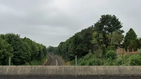 Google A view of a railway line taken from a road bridge. There are trees either side of the line which curves round to the left and out of sight.