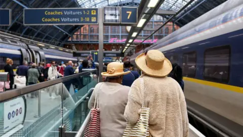 Two women in hats walk alongside a train on a crowded platform in St Pancras station