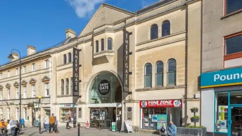 Acorn Property Group The exterior of Emery Gate shopping centre in Chippenham on a sunny day. It is a three story, stone building. Boards outside are advertising the shopping centre. The front of F. Hinds jewellery store and Lloyds Bank is visible on the left while Fone Zone and Poundland are on the right.