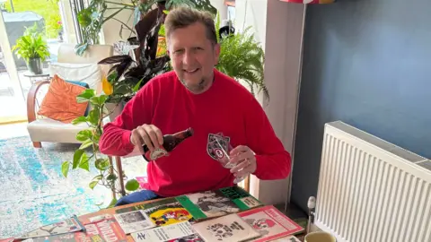 PA Media A man in a vintage red England shirt smiling and pretending to pour a beer in front of a table which is covered in 1966 World Cup memorabilia. 
