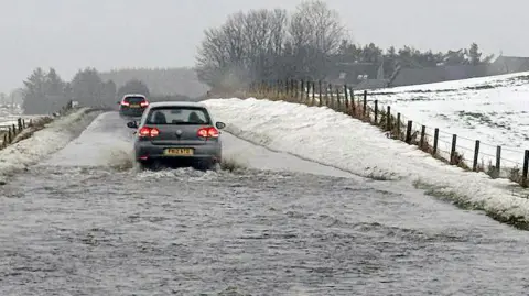 A car drives through floodwater covering a rural road, with snow piled along the roadside and fields under wintry cond