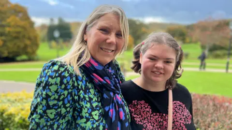 Two people standing outdoors with autumn trees and green lawns in the background, wearing patterned tops and a scarf, with a cloudy sky overhead.