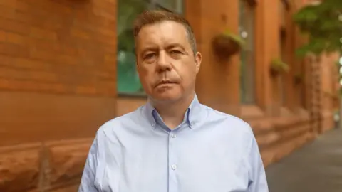 Glyn Roberts, a man with short, dark hair, looks at the camera with a serious expression. He is wearing a blue shirt open at the neck. He is standing on a city centre street in front of a large red brick building. 