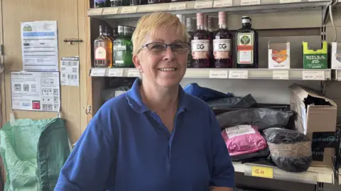 Matt Marvel/BBC A woman inside a shop. She has short blonde hair and is wearing glasses along with a blue polo shirt. Shelves with bottles of alcohol and parcels are behind her.