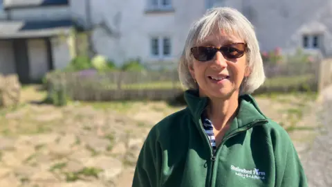 A mid shot of Alison Kohler. She has shoulder length grey hair and is wearing a green jacket, and glasses.