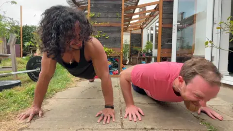 Two men doing push-ups on paving in a garden. The more muscular man is looking at the other man, whose facial expression appears strained.