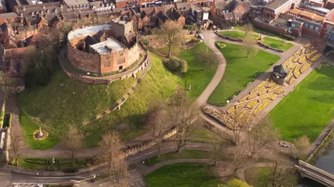 An aerial view of Tamworth Castle, a traditional motte and bailey style castle, sits on top of a mound with more modern buildings to one side and lawned areas to the other.