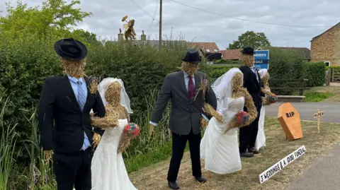 Six life-size figures of three couples in wedding dresses and suits are standing on a grass verge outside a house on the High Street in a rural location, a small wooden coffin with RIP written on the lid next to a sign which says 'Alfie' on it.
A longer white sign stands in front of the display and says 'three weddings and a dog funeral'. Behind the scarecrows is a tall 2.5m (8ft) hedge with a straw model of a dog visible in the middle at the top.