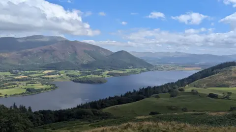 A general view of Bassenthwaite taken from Sale Fell in the Lake District. A large lake dominates the centre of the image. It is surrounded by mountains stretching into the distance, although there is a flatter area sectioned into fields on the far bank. Clumps of dense woodland are dotted around the landscape.