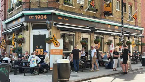 John Drinkall A pub on the corner of a street with orange and white branding. It reads "Boothferry Park" with the letters "fer ark" in bold orange. People are sat on benches outside of the pub, drinking.