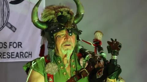 Terry Forster is standing on a stage, holding his bagpipes, and dressed as a Viking. He is wearing a helmet adorned with horns, fur and chainmail. He is wearing a green tunic with metal accessories on it in various Viking patterns. He is lit by a green light.