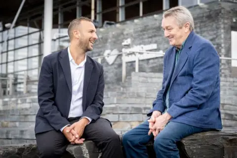 Green Party Zack Polanski and Anthony Slaughter chatting and smiling, sitting together beside the grey steps to the Senedd building in Cardiff Bay.