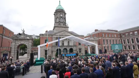 PA Media A large crowd seated in an urban area. In front of the crowd there is an Irish flag and an EU flag. Many of the crowd are wearing suits. The sky is overcast, and the area is surrounded by red brick buildings. 