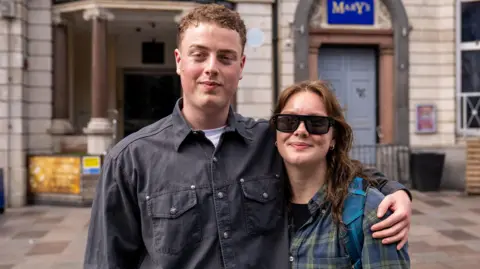 Joe, a man in a charcoal coloured denim shirt with a white tee underneath is stood next to Jess, a woman wearing black, tinted sunglasses and a chequered top. Joe has his hand around Jess; shoulders, and they stand on a street. 