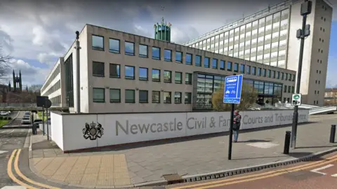 Newcastle Civil and Family Courts and Tribunals Centre is a white three-storey building with winows. Newcastle Civil and Family Courts and Tribunals Centre is written across a white fence alongside the city's Coat of Arms. A person is walkin on the pavement.